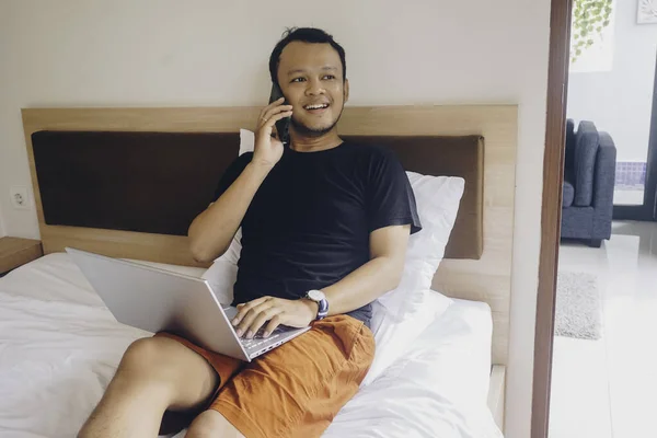 Щасливий Молодий Чоловік Працює Ліжка Тримайте Ноутбук Колінах Розмовляйте Телефону — стокове фото