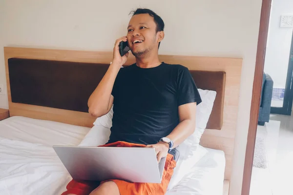 Счастливый Молодой Человек Работающий Постели Держите Ноутбук Коленях Говорите Телефону — стоковое фото