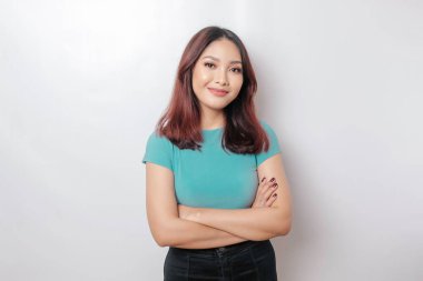 Mavi tişörtlü, kollarını kavuşturup beyaz arka planda izole edilmiş kameraya bakan kendinden emin gülümseyen Asyalı bir kadının portresi.