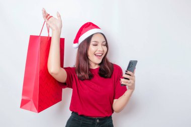 Noel Baba 'nın şapkasını takan mutlu genç bir kadın elinde Noel hediyeleri ve akıllı telefonuyla gülümsüyor.