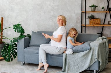 Hamile bir genç kız küçük kızıyla kanepede oturuyor ve telefonda oynuyor. Çocuklarda ve yetişkinlerde aletlere olan bağımlılık kavramı. Yüksek kalite fotoğraf