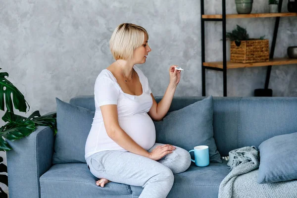 A young blonde pregnant girl sits on a gray sofa in a white T-shirt and gray pants and measures the temperature for the purpose of detecting the disease . High quality photo