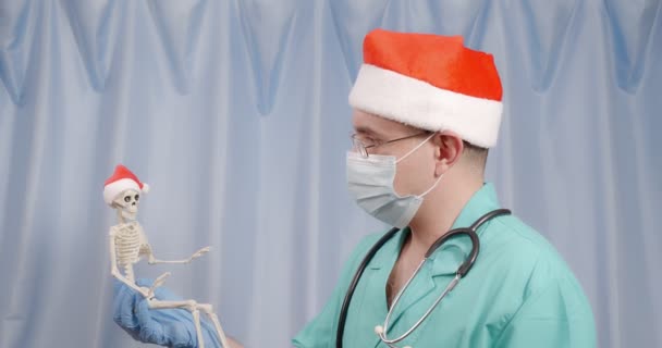 Dokter di topi Santa memegang di tangannya sosok kerangka di topi Santa. — Stok Video