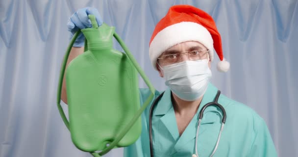 A doctor in a Santa hat holds a large enema in his hands. — Stock Video