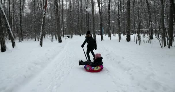 冬の森の屋外で雪のチューブに滑る母と娘 冬の公園で娘とお母さんが遊ぶ — ストック動画