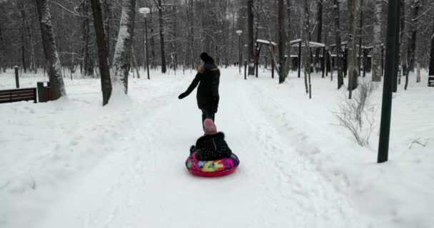 冬の森の屋外で雪のチューブに滑る母と娘 冬の公園で娘とお母さんが遊ぶ — ストック動画