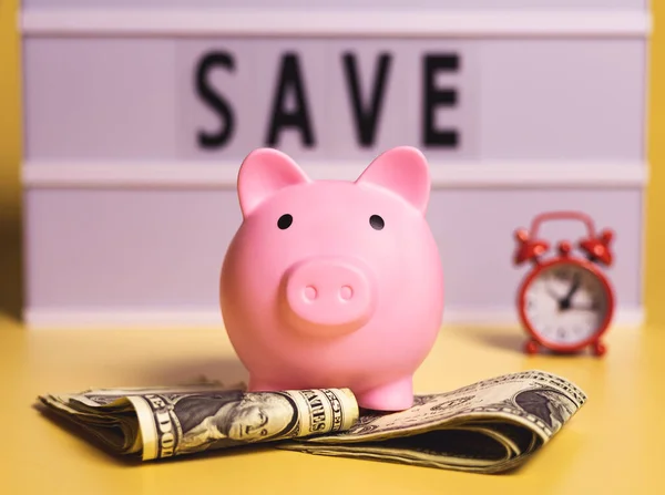 A piggy bank over US dollar banknotes on yellow background. A light box with word save and a small red alarm clock in the image composition.