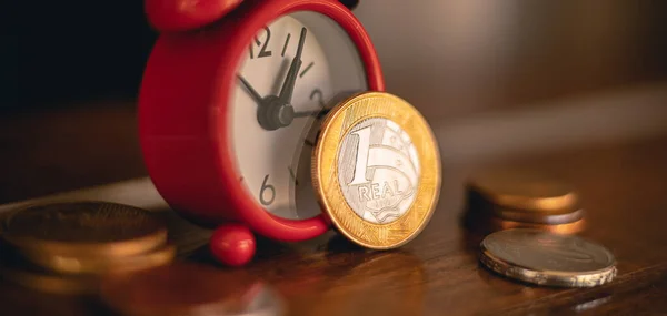Real - BRL, Brazilian currency. Photo with focus on the 1 real coin next to a red clock in a window of a residence. Saving, investment, withdraw, future plans concepts.