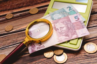 A golden magnifying glass over Brazilian Real banknotes. Coins and a blue calculator in the image composition.