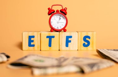ETFS kelimesi stüdyo arka planında tahta küplere yazıldı. Görüntü bileşiminde bir para ve kırmızı bir saat.  