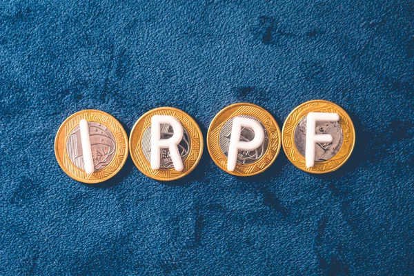 The word The acronym IRPF de Income Tax for Individuals written in white letters on Brazilian coins. Blue background. Brazilian economy concept.