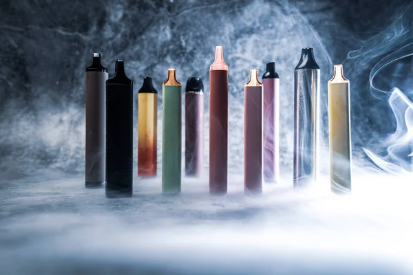 Cigarrillos electrónicos multicolores vapean sobre fondo oscuro con humo Fotos De Stock