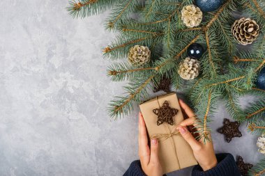 Kadın eli, gri arka planda bir hediye kutusu tutuyor. Kozalaklı ve ladin dallı kompozisyon. Bayramlar için şenlikli arka plan: Noel, Yeni Yıl. Düz yatıyordu.