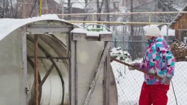 Soğutma, iklim değişikliği. Renkli kışlık elbiseli bir kadın büyük plastik bir kürekle bir çiftlikteki seradan kar temizliyor. Sibirya.