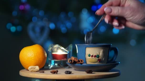 女性はクリスマスツリーの背景に小さなテーブルの上に立ってお茶のカップに砂糖を注ぎ それを攪拌します — ストック動画