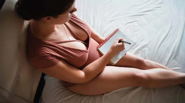 妊娠中の女の子はベッドの上に座り 膝の上にノートで胃をアイロンをかけます 妊産婦病院へのリスト 赤ん坊を待ってる テキスト用の場所 スペースのコピー — ストック写真
