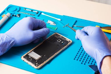 Lastik eldivenli bir zanaatkar tamir için özel bir lastik paspasın üzerindeki cep telefonunu tamir eder ya da ona hizmet eder. İçeriden bak. Cihazı tamir ediyorum. Bozuk akıllı telefon.