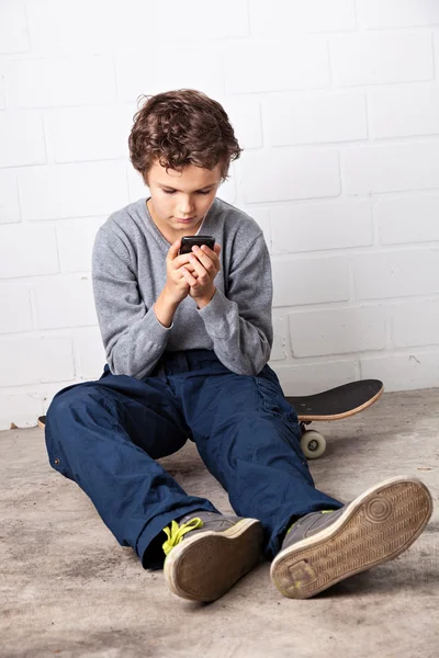 Fajny chłopak siedzi na swojej deskorolce, trzymając smartphone — Zdjęcie stockowe
