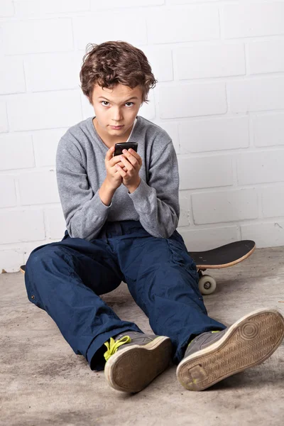 Cooler Junge sitzt auf seinem Skateboard und hält ein Smartphone in der Hand — Stockfoto