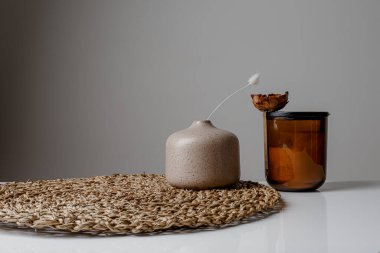 Cozy home interior decor, aromatic candles in glass glowingon white and grey background with natural oil, selective focus of fluffy bunny tail grass, brown jar, lotus flower candle, round matt ceramic