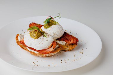 Avocado eggs benedict with pesto sauce, grilled bacon, pepper, cream cheese Philadelphia. Poached eggs on healthy bread toast with hollandaise sauce. Cut egg. The yolk flows out. Micro greens.