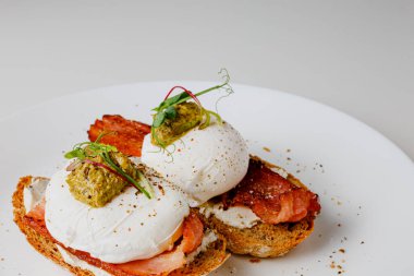 Avocado eggs benedict with pesto sauce, grilled bacon, pepper, cream cheese Philadelphia. Poached eggs on healthy bread toast with hollandaise sauce. Cut egg. The yolk flows out. Micro greens.