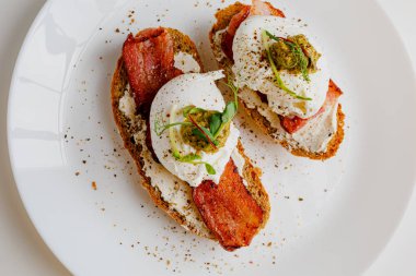 Avocado eggs benedict with pesto sauce, grilled bacon, pepper, cream cheese Philadelphia. Poached eggs on healthy bread toast with hollandaise sauce. Cut egg. The yolk flows out. Micro greens.