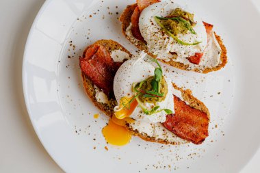 Avocado eggs benedict with pesto sauce, grilled bacon, pepper, cream cheese Philadelphia. Poached eggs on healthy bread toast with hollandaise sauce. Cut egg. The yolk flows out. Micro greens.