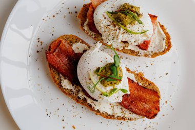 Avocado eggs benedict with pesto sauce, grilled bacon, pepper, cream cheese Philadelphia. Poached eggs on healthy bread toast with hollandaise sauce. Cut egg. The yolk flows out. Micro greens.