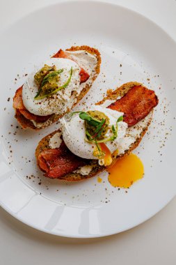 Avocado eggs benedict with pesto sauce, grilled bacon, pepper, cream cheese Philadelphia. Poached eggs on healthy bread toast with hollandaise sauce. Cut egg. The yolk flows out. Micro greens.