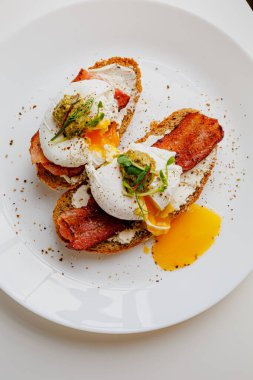 Avocado eggs benedict with pesto sauce, grilled bacon, pepper, cream cheese Philadelphia. Poached eggs on healthy bread toast with hollandaise sauce. Cut egg. The yolk flows out. Micro greens.