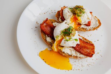 Avocado eggs benedict with pesto sauce, grilled bacon, pepper, cream cheese Philadelphia. Poached eggs on healthy bread toast with hollandaise sauce. Cut egg. The yolk flows out. Micro greens.