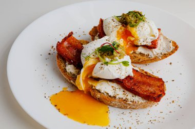 Avocado eggs benedict with pesto sauce, grilled bacon, pepper, cream cheese Philadelphia. Poached eggs on healthy bread toast with hollandaise sauce. Cut egg. The yolk flows out. Micro greens.