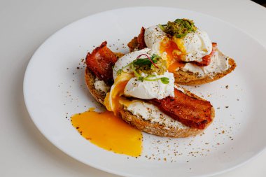 Avocado eggs benedict with pesto sauce, grilled bacon, pepper, cream cheese Philadelphia. Poached eggs on healthy bread toast with hollandaise sauce. Cut egg. The yolk flows out. Micro greens.