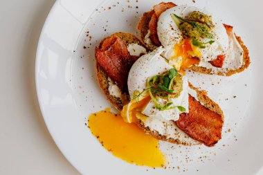 Avocado eggs benedict with pesto sauce, grilled bacon, pepper, cream cheese Philadelphia. Poached eggs on healthy bread toast with hollandaise sauce. Cut egg. The yolk flows out. Micro greens.