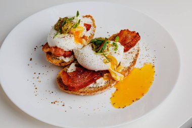 Avocado eggs benedict with pesto sauce, grilled bacon, pepper, cream cheese Philadelphia. Poached eggs on healthy bread toast with hollandaise sauce. Cut egg. The yolk flows out. Micro greens.