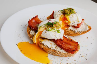 Avocado eggs benedict with pesto sauce, grilled bacon, pepper, cream cheese Philadelphia. Poached eggs on healthy bread toast with hollandaise sauce. Cut egg. The yolk flows out. Micro greens.