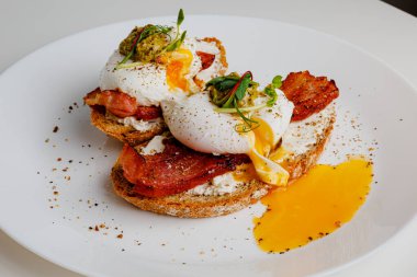 Avocado eggs benedict with pesto sauce, grilled bacon, pepper, cream cheese Philadelphia. Poached eggs on healthy bread toast with hollandaise sauce. Cut egg. The yolk flows out. Micro greens.