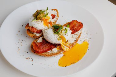 Avocado eggs benedict with pesto sauce, grilled bacon, pepper, cream cheese Philadelphia. Poached eggs on healthy bread toast with hollandaise sauce. Cut egg. The yolk flows out. Micro greens.