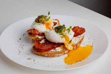 Avocado eggs benedict with pesto sauce, grilled bacon, pepper, cream cheese Philadelphia. Poached eggs on healthy bread toast with hollandaise sauce. Cut egg. The yolk flows out. Micro greens.