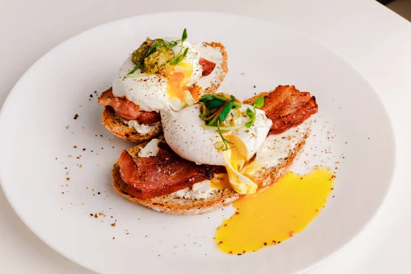 Avocado eggs benedict with pesto sauce, grilled bacon, pepper, cream cheese Philadelphia. Poached eggs on healthy bread toast with hollandaise sauce. Cut egg. The yolk flows out. Micro greens.