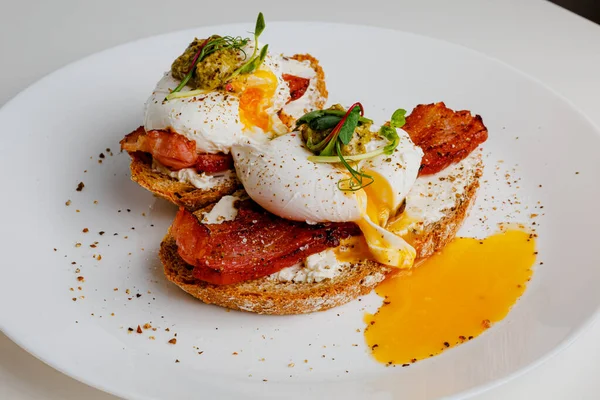 Avocado eggs benedict with pesto sauce, grilled bacon, pepper, cream cheese Philadelphia. Poached eggs on healthy bread toast with hollandaise sauce. Cut egg. The yolk flows out. Micro greens.