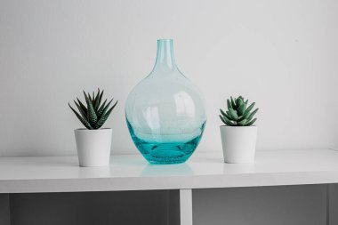 Decorative artificial ceramic and metal cactus, succulent and cactuses on white wall background. Light blue and mint glass vase. Home decoration. Side view. Copy space.
