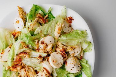 Traditional Caesar salad in a white bowl plate on a white table with green leaves and a sauce boat and parmesan and mozzarella cheese balls. Selected focus.