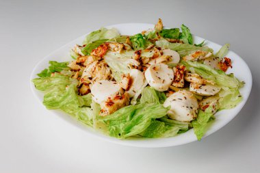 Traditional Caesar salad in a white bowl plate on a white table with green leaves and a sauce boat and parmesan and mozzarella cheese balls. Selected focus.