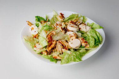 Traditional Caesar salad in a white bowl plate on a white table with green leaves and a sauce boat and parmesan and mozzarella cheese balls. Selected focus.