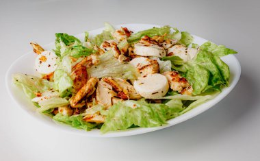 Traditional Caesar salad in a white bowl plate on a white table with green leaves and a sauce boat and parmesan and mozzarella cheese balls. Selected focus.