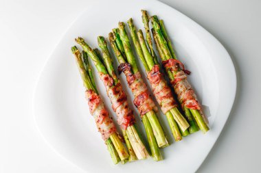 Grilled green asparagus wrapped with bacon. Asparagus baked with bacon and spices. Healthy food. Top view. Free space for your text. Bacon wrapped asparagus on white plate.