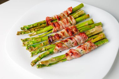 Grilled green asparagus wrapped with bacon. Asparagus baked with bacon and spices. Healthy food. Top view. Free space for your text. Bacon wrapped asparagus on white plate.