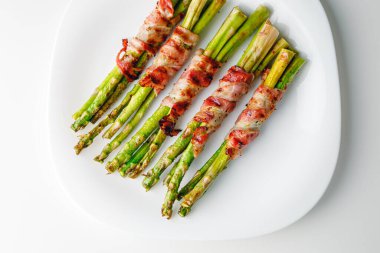 Grilled green asparagus wrapped with bacon. Asparagus baked with bacon and spices. Healthy food. Top view. Free space for your text. Bacon wrapped asparagus on white plate.